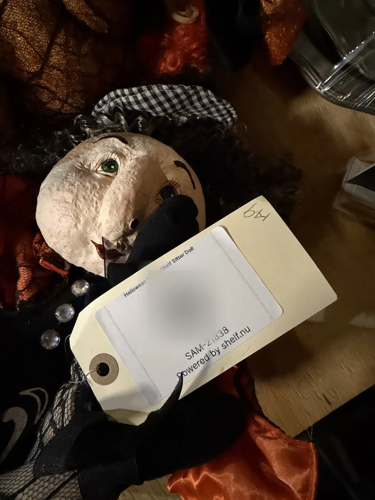 A Shelf label on a manilla hang tag for a Halloween witch shelf sitter doll, showing the item name, a blurred QR code and the SAM ID