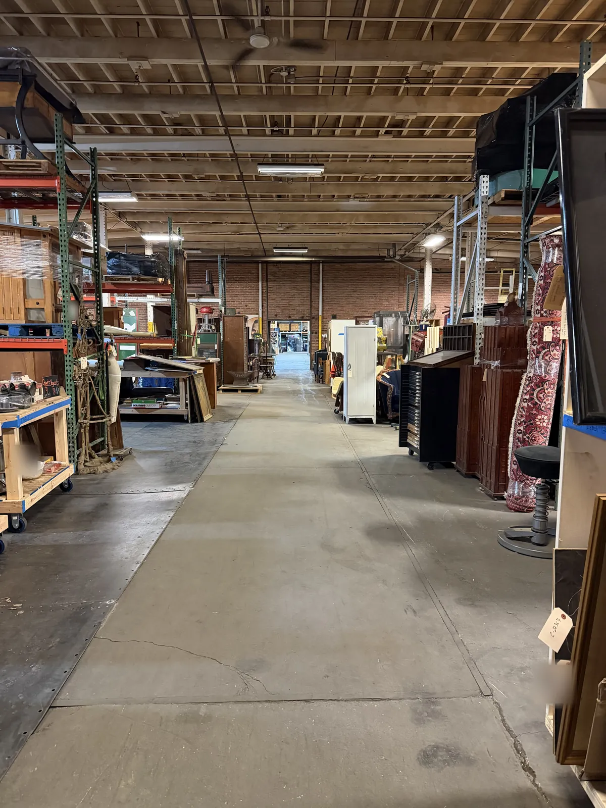 A long warehouse aisle at Industrial Artifacts, pallet racking and furniture on both sides and a rolling table on the left